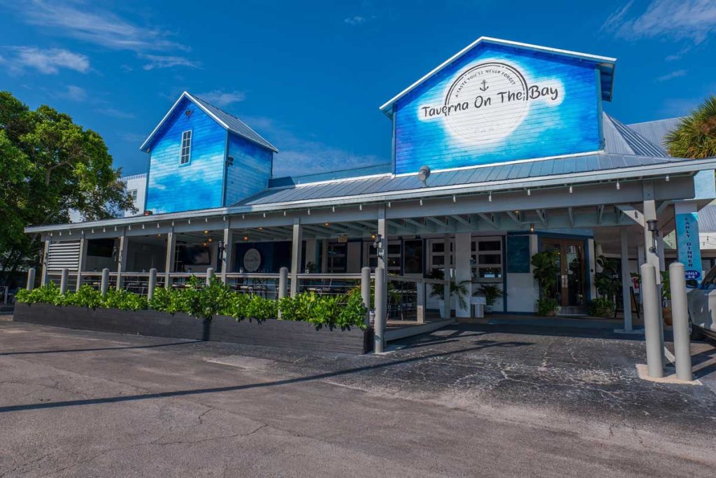 A photo of a blue and white restaurant with an outdoor eating area surrounded by plants; this is Taverna on the Bay in St. Pete Beach.